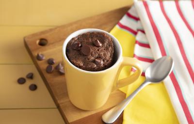 Fotografia em tons de amarelo em uma bancada de madeira amarela, um pano listrado vermelho e amarelo e uma tábua de madeira ao centro da foto com uma caneca amarela com o bolo de caneca de chocolate apoiada sobre ela.