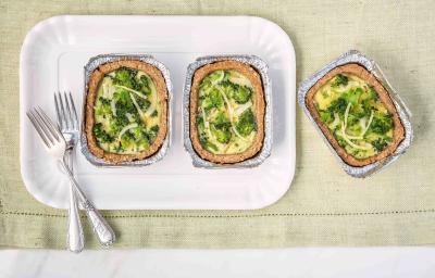 Fotografia em tons de branco, verde e dourado de uma bancada vista de cima mostrando refratário branco com duas porções de quiche com brócolis e dois garfos, com mais uma porção ao lado, tudo sobre guardanapo em tom de branco.