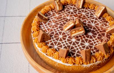 Fotografia em tons de caramelo e branco um um prato redondo de madeira ao centro. Em cima do prato existe uma torta em tons de caramelo e chocolate com alguns pedaços de chocolate no topo