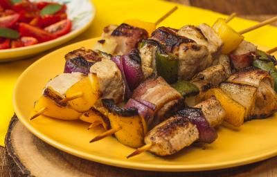 Foto bem aproximada de um prato amarelo sobre o qual há quatro espetinhos montados com carne de porco, cebolas roxa e tradicional e pimentões verdes e amarelos. Ao fundo há um prato branco com uma salada de tomates.