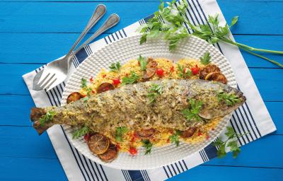 Fotografia em tons de azul e branco de uma bancada de madeira azul com um paninho branco e azul listrado, sobre ele um prato branco oval com um peixe assada e farofa. Ao fundo um garfo e um ramo de salsinha.