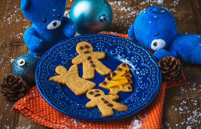 Fotografia em tons de laranja e azul em uma bancada de madeira, um paninho laranja, um prato azul com os biscoitinhos natalinos em cima dele. Ao lado, decoração e enfeites de natal.