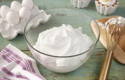 Fotografia em tons de verde em uma bancada de madeira verde, um pano branco com listras roxas, um recipiente de vidro com o merengue dentro dele. Ao fundo, potinhos com docinhos cobertos com merengue, ovos, tábua de madeira com um fouet.