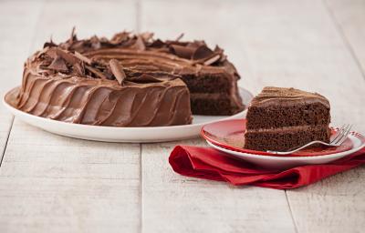 Fotografia em tons chocolate e branco, ao centro prato com bolo de chocolate fatiado, fatia ao lado sobre pratos vermelho e branco com um garfo e sobre guardanapo vermelho, tudo sobre bancada de madeira em pátina.