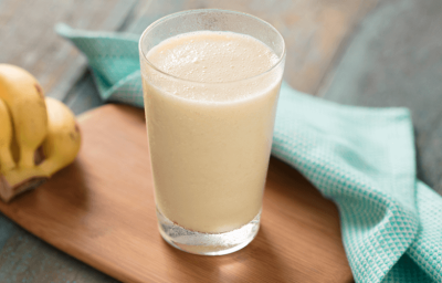 Fotografia em tons de azul e amarelo em uma bancada de madeira escura, uma tábua de madeira, um pano de pano azul com bolinhas, um cacho de bananas e um copo de vidro alto com a vitamina de banana com maracujá e coco.