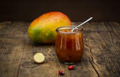 Fotografia de um chutney de manga e laranja temperado dentro de um recipiente de vidro pequeno, como se fosse um copo. Apoiado no chutney, uma colher de chá, e ao fundo, uma manga inteira com casca, sobre uma mesa de madeira.