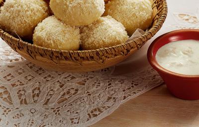 Fotografia em tons de branco, marrom, pardo e vermelho de uma bancada vista de cima. Um pano de renda branco e por cima um recipiente cor pardo e redondo com pães bolinha, ao lado um potinho vermelho redondo com creme branco.