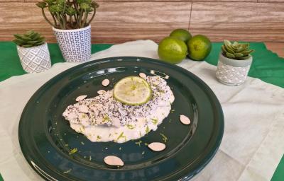 Foto em tons de branco da receita de tapioca servida com uma rodela de limão por cima, sobre um prato preto em cima de uma toalha de linho com limões e vasinhos de planta ao lado