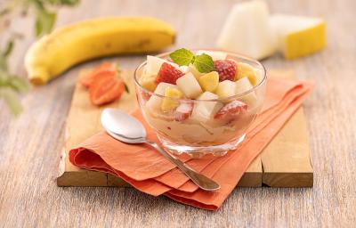 Foto da receita de salada de frutas servida em uma taça de vidro decorada com uma folha de hortelã, sobre um pano laranja com uma colher prateada ao lado. Ao fundo, há morangos, uma banana e fatia de melão