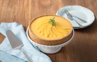 fotografia em tons de marrom, branco e amarelo de uma bancada marrom vista de frente, contém uma bailarina para servir sobremesa branca com uma torta por cima. Ao lado esquerdo contém um prato redondo branco e do lado direito um pano azul com uma espátula