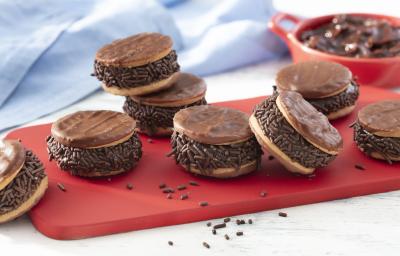 Fotografia em tons de vermelho em uma bancada de madeira clara, um pano azul, uma tábua vermelha com vários sanduíches de biscoito calipso recheados com brigadeiro de pão de mel. Ao fundo, um potinho vermelho com o brigadeiro dentro.