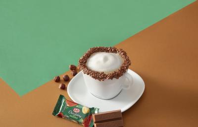 Foto da receita de cappuccino kit kat servida em uma xícara branca com um kit kat avelã ao lado em fundo marrom e verde