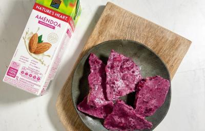 Foto em tons de roxo da receita de barrinha proteica servida em algumas porções em um prato cinza escuro sobre uma tábua de madeira com uma embalagem de bebida vegetal de amêndoas nature's heart ao lado