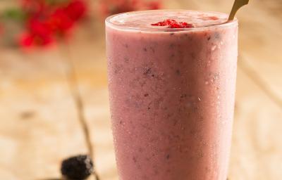 Fotografia em tons de rosa em uma mesa de madeira com um copo de vidro ao centro com o shake de frutas vermelhas dentro. Morangos e mirtilos espalhados pela mesa.