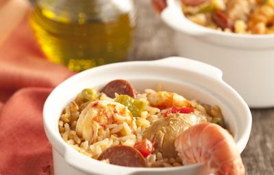 fotografia em tons de cinza e branco de uma bancada vista de frente, contém dois potinhos brancos e redondos com arroz, pedaços de carne suína e frutos do mar. Ao lado um pano laranja com dois garfos para servir e uma jarra de azeite