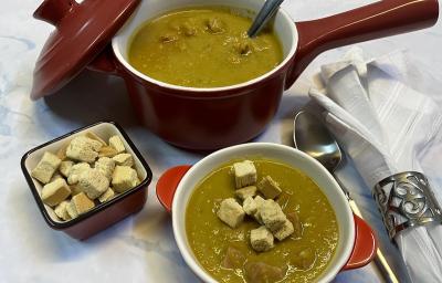 \foto em tons de verde da receita de creme de ervilha servida em uma panelinha marrom com uma colher de servir prateada dentro, à frente há um bowl laranja e branco com uma porção do creme e ao lado há crouttons e uma colher prateada