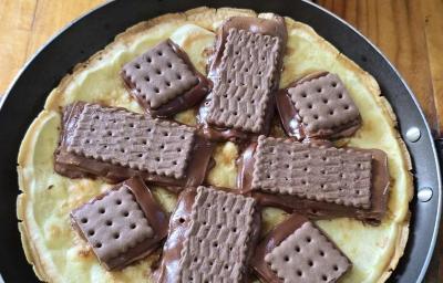 Fotografia em tons de marrom e vermelho com um prato preto ao centro. Em cima do prato tem uma pizza feita de massa de ovo e tapioca. Por cima da massa existe vários biscoitos Choco Trio derretidos.