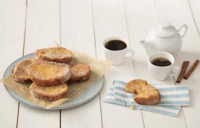 Fotografia em tons de dourado e branco, com rabanadas fritas sobre papel manteiga e prato azul, ao lado serviço de café, paus de canela e rabanada sobre guardanapo listrado em azul e branco, tudo sobre bancada em tom pastel.