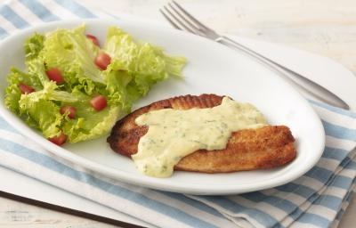 Fotografia em tons de azul em uma bancada de madeira clara com um pano listrado azul claro e um prato oval branco com o filé de pescada com molho de limão e mostrada e salada de alface com tomates.