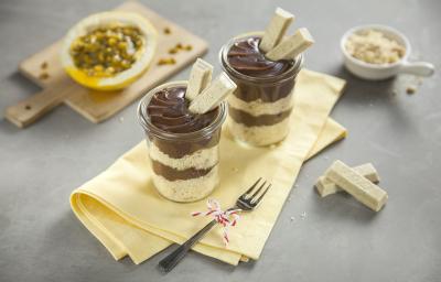 Fotografia em tons de amarelo em uma bancada cinza com um pano amarelo ao centro com dois potinhos de vidro com o bolo de pote de brigadeiro com dois KitKat de Passion Fruit decorando o pote. Ao fundo, um maracujá cortado ao meio.