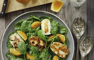 Fotografia vista de cima de uma salada de agrião, frango defumado e gomos de laranja, que está em um prato fundo cinza, sobre uma mesa de madeira. Ao lado, uma tábua de madeira de tom claro, com uma laranja inteira, cascas da fruta e uma faca apoiada.