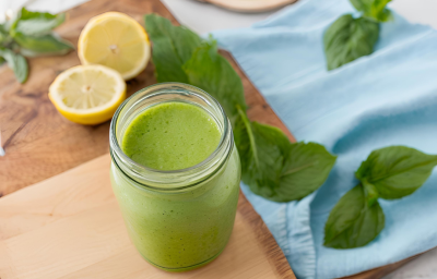 Fotografia de um copo de vidro decorativo com suco detox de cor verde feito com couve, maçã verde, pepino, gengibre e nutren. O copo está sobre uma tábua de madeira, ao lado de folhas de hortelã e metades de limão siciliano sobre a mesa.