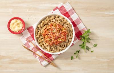 Fotografia em tons de vermelho em uma bancada de madeira com um pano de xadrez vermelho, em cima um prato fundo redondo com o macarrão integral com molho de frango dentro. Ao lado, um pegador de macarrão e um potinho vermelho com queijo ralado.