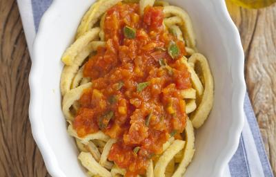 fotografia em tons de marrom, branco, bege e vermelho de uma bancada marrom vista de cima, ao centro um pano listrado em tons de azul e branco com um recipiente branco oval com macarrão e molho de tomate por cima