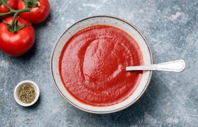 fotografia em tons de cinza e vermelho de uma bancada cinza vista de cima. Ao centro um recipiente redondo com sopa de tomate e um colher para servir, e ao lado esquerdo contém tomates.