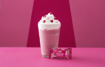 Foto da receita de Frappé. Observa-se um fundo rosa com um copo alto decorado de chantily e confeitos de coração com um kit kat de morango na frente.