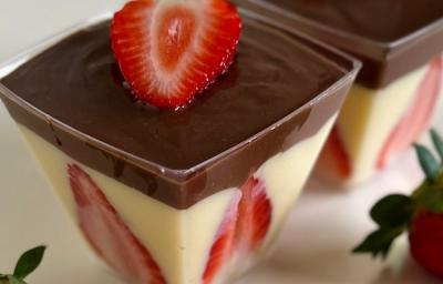 Fotografia de dois potes quadrados transparentes com bombom de morango com ganache de chocolate, morangos decorando a parede do copinho junto ao creme branco, e tem uma fatia por cima. Os potinhos estão sobre uma mesa branca, com morangos ao redor. Fotografia de dois potes quadrados transparentes com bombom de morango com ganache de chocolate, morangos decorando a parede do copinho junto ao creme branco, e tem uma fatia por cima. Os potinhos estão sobre uma mesa branca, com morangos ao redor.