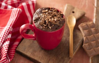 Fotografia em tons de vermelho de uma bancada de madeira, ao centro uma tábua de madeira com uma caneca vermelha e um bolo de chocolate com raspas de chocolate dentro. Ao lado uma pano vermelho com listras brancas e uma colher de madeira.