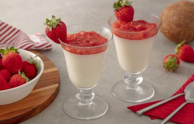 Foto da Receita de Taças de coco com purê de morango. Observa-se duas taças de vidro com o mousse e o purê com morangos decorando a borda da taça. Como decoração, vemos morangos ao lado direito em um potinho e um coco fresco ao lado direito