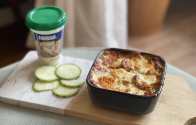 Fotografia em tons de marrom e verde em uma bancada de madeira, com uma tábua de madeira e em cima um pote quadrado com a lasanha de abobrinha com frango e requeijão light. Ao lado, um pote de requeijão nestlé.