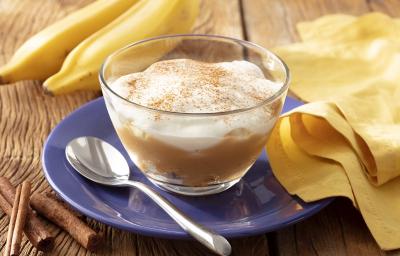 Fotografia em tons de branco em uma bancada de madeira de cor marrom. Ao centro, um prato azul contendo um recipiente com a banoffee. Ao lado, há uma colher, um pano amarelo e ao fundo, 2 bananas.