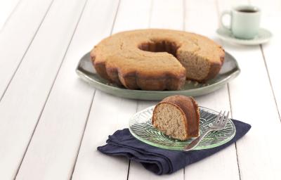 Fotografia em tons de verde em uma bancada de madeira branca, um paninho verde, um prato redondo grande com o bolo de amêndoas e aveia com um pedaço cortado e um pratinho pequeno com a fatia do bolo.