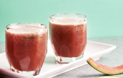 Fotografia em tons de rosa e vermelho em uma bancada de madeira clara com um recipiente branco retangular com o suco de melancia e menta. Ao lado, fatias de melancia e uma melancia inteira.