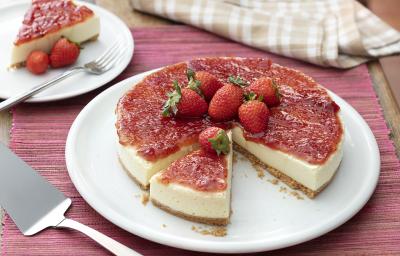 Fotografia em tons de vermelho, em uma bancada de madeira de cor marrom. No centro, uma toalha vermelha e em cima um prato redondo branco contendo o cheesecake. Ao fundo, um pires branco, contendo uma fatia do cheesecake com 2 morangos e um garfo ao lado.