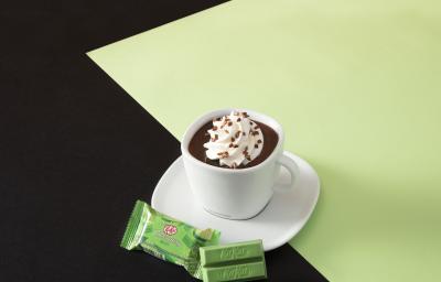 Foto da receita de Chocolate Quente Cremoso. Observa-se uma xícara com pires brancos de porcelana com chocolate dentro, creme por cima e granulado polvilhado. O fundo é metade marrom e metade verde, servido com um kit kat de menta.