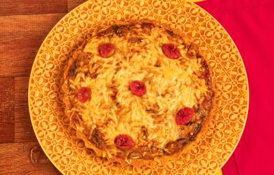 Fotografia mostra uma quiche com queijo gratinado e pedaços de tomate sobre um prato amarelo. Ao fundo, uma mesa de madeira e um pano vermelho.