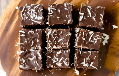 A foto mostra quadradinhos de cocada de chocolate densos e brilhantes em uma travessa rústica de madeira. A cocada tem uma cor marrom profunda, com pedaços de coco visíveis.