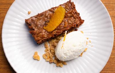 Fotografia de um prato branco raso com um pedaço de bolo de chocolate cremoso com crocante de nozes, e por cima, chantilly de laranja. O prato está sobre uma bancada de madeira clara.