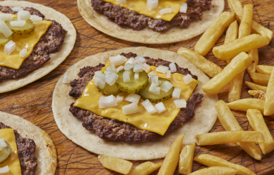 Fotografia mostra uma porção de tacos abertos, sob uma mesa de madeira, com recheio de carne bovina, queijo, cebola, alface e picles para finalizar.