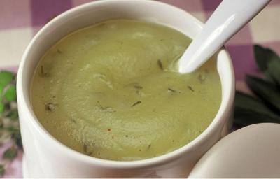 Fotografia em tons de verde em uma mesa com uma toalha roxa xadrez e uma xícara branca aproximada com a geleia verde de ervas.