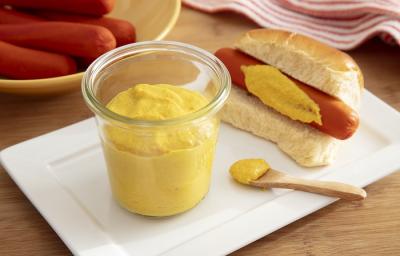 Foto da receita de mostarda caseira com maggi. Observa-se um potinho transparente com a mostarda e, ao lado, um hot dog com o molho passado na salsicha. Também há uma colherinha de madeira com um pouco do molho.