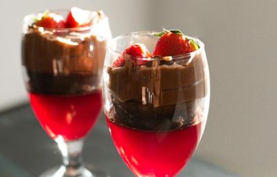 Fotografia em tons de vermelho em uma bancada de madeira cinza escura com duas taças de vidro com o doce de morango com uma camada de brownie de chocolate, outra com mousse de chocolate e finalizando com morangos para decorar.