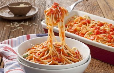 Fotografia em tons de vermelho em uma bancada de madeira com um pano branco com listras vermelhas e azuis, um prato fundo branco com o macarrão ao molho sugo sendo puxado por um garfo. Ao fundo, um recipiente com macarrão e um potinho de queijo ralado.