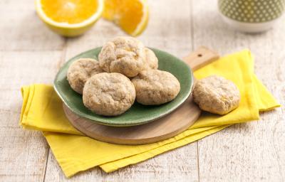 Fotografia em tons de amarelo em uma bancada de madeira clara, um pano amarelo, uma tábua de madeira com um prato verde redondo com alguns biscoitinhos de banana com laranja em cima dele. Ao fundo, uma laranja cortada ao meio e uma xícara verde.