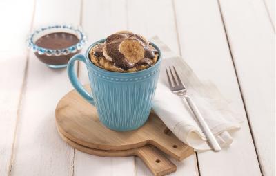 Foto da receita de bolo de caneca com cacau e banana em uma caneca azul, sobre tábuas de madeira, com um guardanapo de pano branco e um garfo sobre ele.