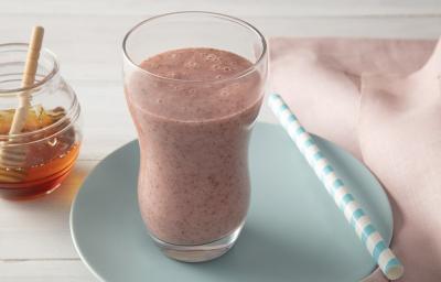 Foto da receita de smoothie iogurte molico com morango e chia servida em um copo largo sobre um pratinho azul com um canudo listrado ao lado junto deum paninho rosa. Ao fundo há um vidro de mel